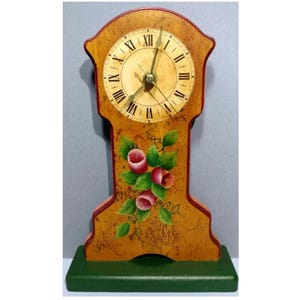 Może przedstawiać: Drewniany zegar z złotą tarczą i cyframi rzymskimi. Zegar jest pomalowany kwiatowym wzorem z różowymi kwiatami i zielonymi liśćmi. Zegar ma zieloną podstawę.