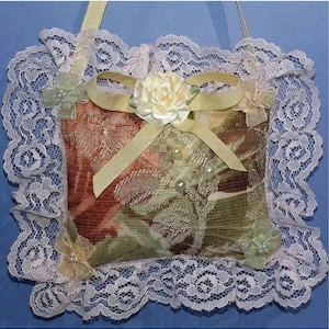 Peut inclure: Un petit coussin carré décoratif avec un imprimé floral dans des tons de vert, marron et crème. Le coussin est bordé de dentelle blanche et a un nœud en satin jaune avec une fleur crème au centre.