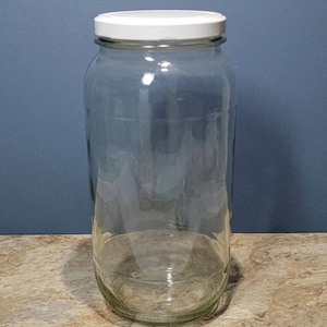 May include: A clear glass jar with a white lid. The jar is tall and cylindrical, with a slightly rounded bottom. It is empty and ready to be filled.