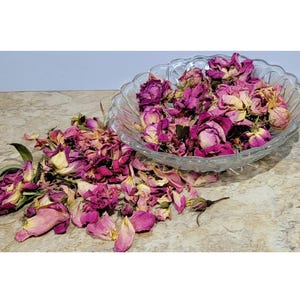 Könnte beinhalten: Eine Glasschüssel gefüllt mit getrockneten rosa Rosenblüten und -knospen. Die Schüssel steht auf einer hellbraunen Arbeitsplatte. Einige Blütenblätter und Knospen sind aus der Schüssel gefallen.