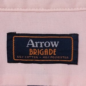 K&ouml;nnte beinhalten: Ein rosa, tailliertes Hemd mit einem schwarzen Etikett mit der Aufschrift "Arrow Brigade 60% Baumwolle - 40% Polyester" und einem kleineren schwarzen Etikett mit dem Text "14 1/2 32/33".