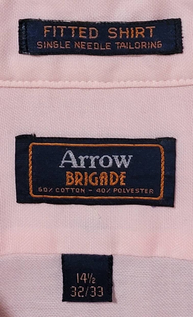 K&ouml;nnte beinhalten: Ein rosa, tailliertes Hemd mit einem schwarzen Etikett mit der Aufschrift "Arrow Brigade 60% Baumwolle - 40% Polyester" und einem kleineren schwarzen Etikett mit dem Text "14 1/2 32/33".