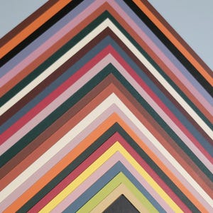 May include: A collection of colorful paper mats arranged in a zig-zag pattern. The mats are in various shades, including black, brown, orange, pink, and green. The background is a light blue. The mats are likely for framing artwork or photographs.