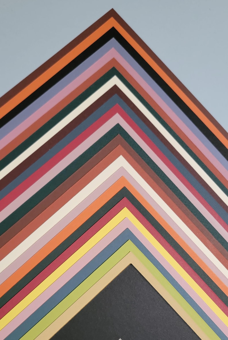May include: A collection of colorful paper mats arranged in a zig-zag pattern. The mats are in various shades, including black, brown, orange, pink, and green. The background is a light blue. The mats are likely for framing artwork or photographs.