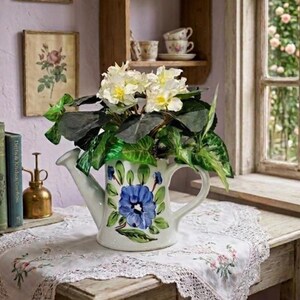 Può includere: Un annaffiatoio decorativo in ceramica bianca con un disegno di fiore blu dipinto a mano e foglie verdi. Il vaso contiene una rigogliosa composizione di fiori bianchi e fogliame verde scuro, creando un elemento botanico decorativo.