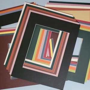 May include: A collection of colorful picture mats in various sizes and colors, including black, brown, green, and white. The mats are arranged in a layered pattern, with rectangular openings for displaying artwork or photos. The mats are for framing.