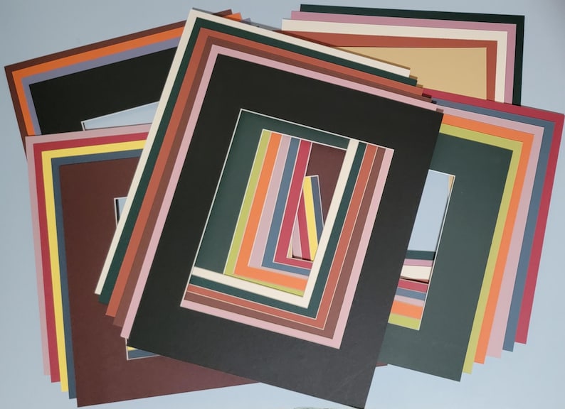 May include: A collection of colorful picture mats in various sizes and colors, including black, brown, green, and white. The mats are arranged in a layered pattern, with rectangular openings for displaying artwork or photos. The mats are for framing.