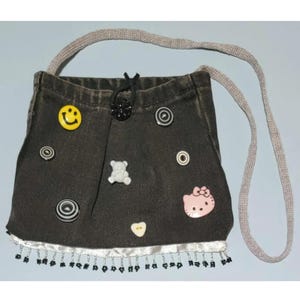 Peut inclure: Petit sac bandoulière en jean noir avec une sangle grise. Le sac est orné de divers embellissements, dont un bouton smiley jaune, un ours en peluche blanc et un Hello Kitty rose. Le bord inférieur a une garniture blanche avec des perles noires.