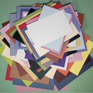 May include: A colorful assortment of layered cardstock paper in various sizes and hues, including white, blue, pink, green, and brown. The paper is arranged in a pile, with a green background.