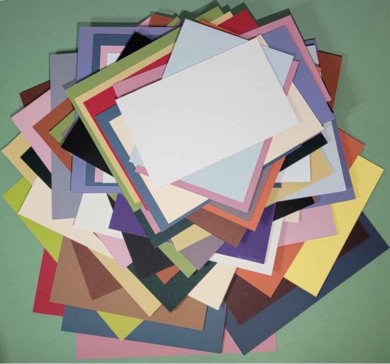 May include: A colorful assortment of layered cardstock paper in various sizes and hues, including white, blue, pink, green, and brown. The paper is arranged in a pile, with a green background.