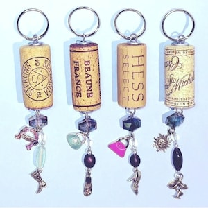 May include: Four keychains made from wine corks with different labels. Each keychain has a charm attached to it, including a horseshoe, a purse, a sun, and a shoe.