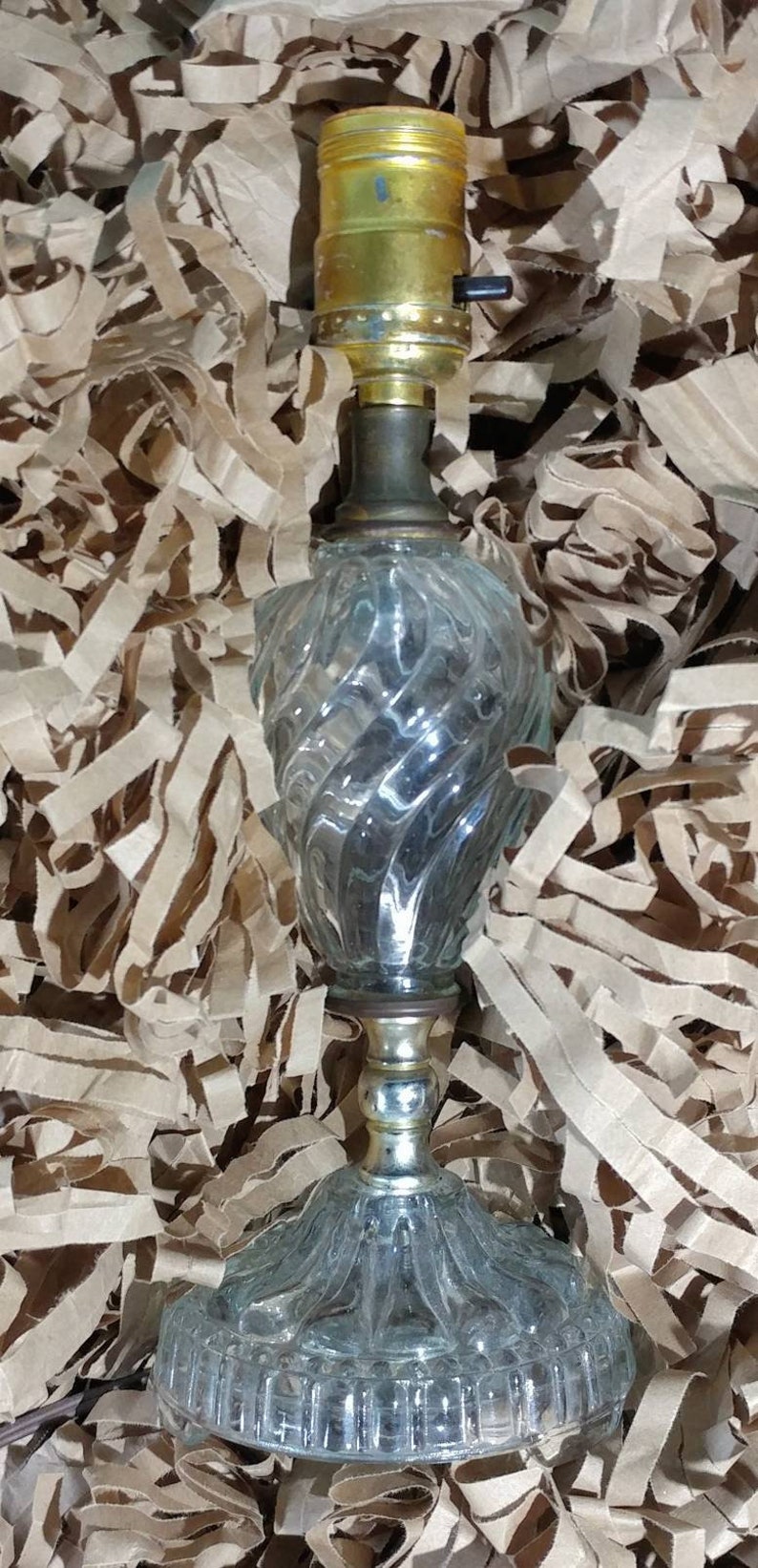 May include: A clear glass table lamp with a gold metal base and a twist design. The lamp has a cord and a switch on the side.