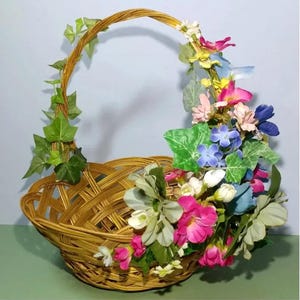 May include: A woven wicker basket with a handle decorated with artificial flowers and greenery. The flowers are pink, blue, white, and yellow.