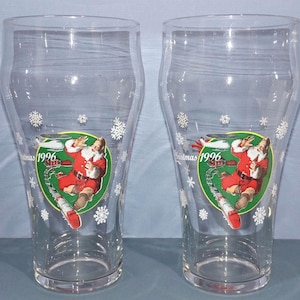 May include: Two clear glass pint glasses with a Christmas theme. Each glass features a circular graphic of Santa Claus on skis, with the text "Christmas 1996". White snowflakes are scattered around the glass.