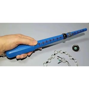 May include: Blue plastic ring sizer with a white ruler marked in sizes. The sizer is held in a hand. A silver chain with white and black beads is in the background.