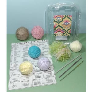 Puede incluir: Un folleto de instrucciones de punto de crochet con ovillos de lana de colores, dos agujas de crochet y un pequeño recipiente de plástico con una tela estampada en su interior.