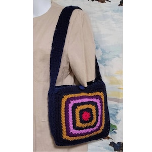 Puede incluir: Un bolso de hombro de crochet con una correa azul marino y un diseño cuadrado colorido. El cuadrado presenta un círculo rojo en el centro rodeado de anillos concéntricos de morado, rosa, dorado y marrón.