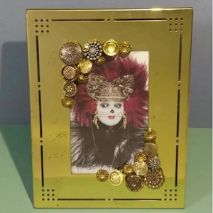 Puede incluir: Un marco de fotos de color dorado con un borde decorativo de botones y una foto de una persona que lleva una máscara de carnaval. La máscara es roja, dorada y negra con plumas.