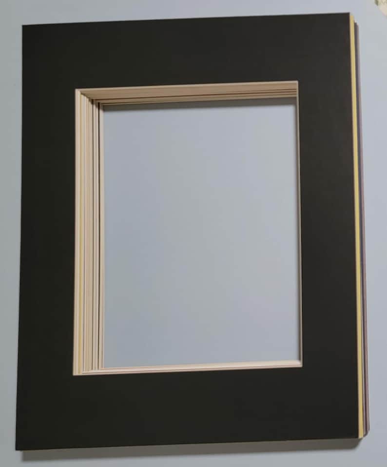 May include: A rectangular black picture frame mat with a light beige inner border. The mat has a large rectangular opening in the center. The edges of the mat are layered with a light beige and yellow accent.