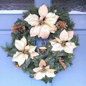 Puede incluir: Una corona navideña festiva con flores de pascua color crema, agujas de pino y piñas. La corona está adornada con detalles dorados y se muestra en una puerta azul. Un llamador decorativo es visible en el centro.