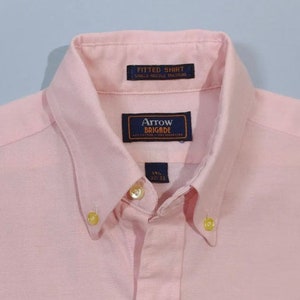 K&ouml;nnte beinhalten: Ein rosafarbenes, lang&auml;rmliges Hemd mit Kn&ouml;pfen und einem blauen Etikett mit der Aufschrift "Arrow Brigade" sowie einem kleineren Etikett mit der Aufschrift "Fitted Shirt Single Needle Tailoring".