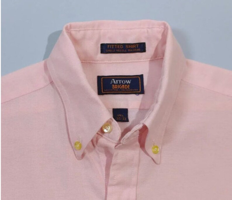K&ouml;nnte beinhalten: Ein rosafarbenes, lang&auml;rmliges Hemd mit Kn&ouml;pfen und einem blauen Etikett mit der Aufschrift "Arrow Brigade" sowie einem kleineren Etikett mit der Aufschrift "Fitted Shirt Single Needle Tailoring".