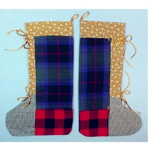 Może przedstawiać: Dwie ręcznie robione świąteczne skarpety z patchworkowym wzorem. Skarpety są wykonane z różnych tkanin, w tym kraty, czerwono-czarnej kraty w kratę i wzoru kwiatowego. Skarpety mają złote wstążki.