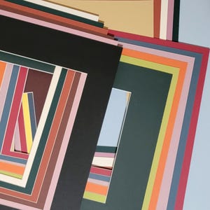 May include: A collection of colorful picture mats in various sizes and hues, including black, green, pink, and orange. The mats are stacked, showcasing a range of rectangular and square openings for artwork or photos.