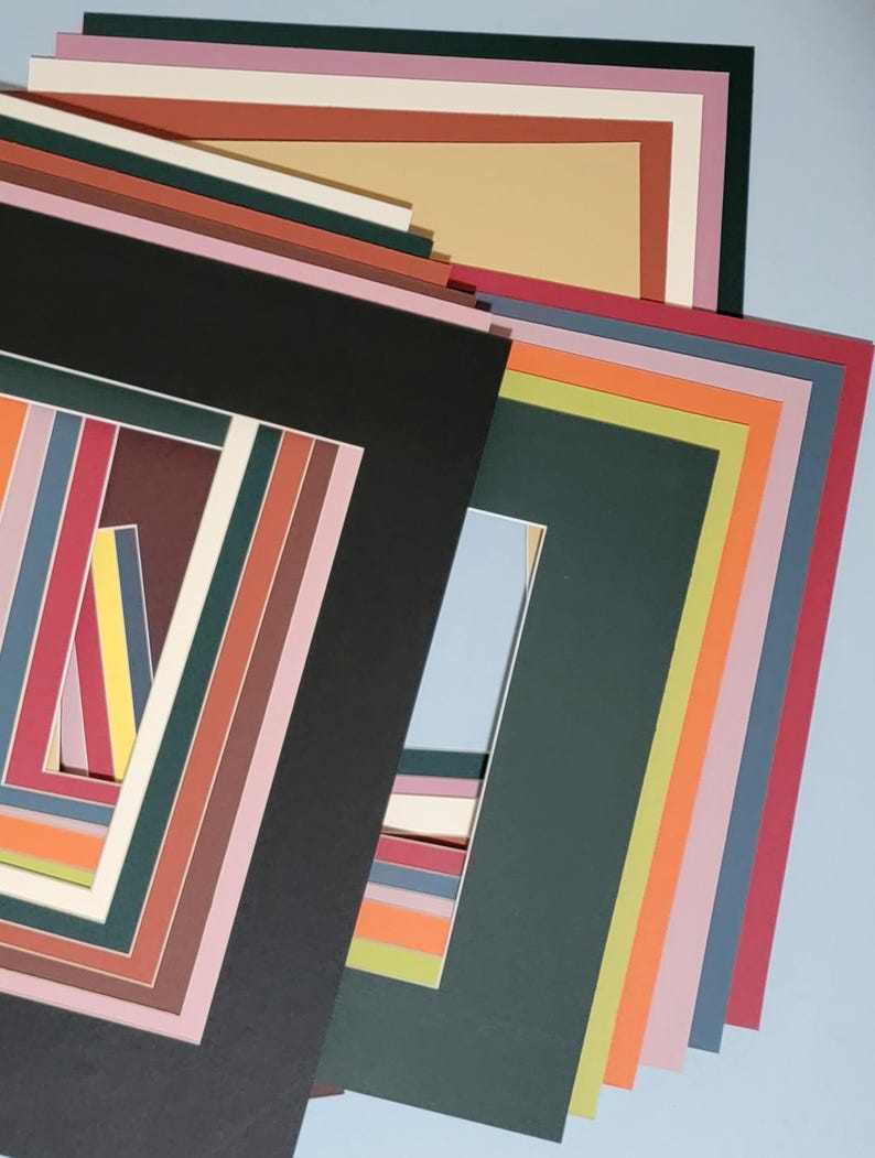 May include: A collection of colorful picture mats in various sizes and hues, including black, green, pink, and orange. The mats are stacked, showcasing a range of rectangular and square openings for artwork or photos.