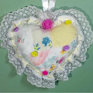 May include: A heart-shaped patchwork cushion with a white lace trim. The cushion is made of various floral fabrics in shades of yellow, pink, and blue. It is decorated with small, colourful buttons and a pink fabric flower.