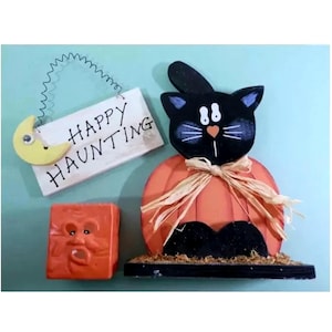 Könnte beinhalten: Halloween-Dekorationen aus Holz. Eine schwarze Katze mit einem Kürbiskörper und einem Schild mit der Aufschrift "Happy Haunting" mit einem gelben Mond. Außerdem gibt es ein kleines orangefarbenes Quadrat mit einem Gesicht.