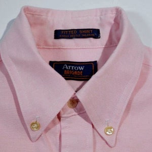 K&ouml;nnte beinhalten: Ein rosa Hemd mit Kn&ouml;pfen und wei&szlig;em Kragen. Das Hemd hat zwei goldene Kn&ouml;pfe und ein Etikett mit der Aufschrift "Arrow Brigade" und "Fitted Shirt Single Needle Tailoring".