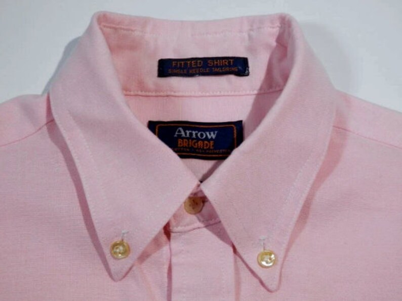 K&ouml;nnte beinhalten: Ein rosa Hemd mit Kn&ouml;pfen und wei&szlig;em Kragen. Das Hemd hat zwei goldene Kn&ouml;pfe und ein Etikett mit der Aufschrift "Arrow Brigade" und "Fitted Shirt Single Needle Tailoring".