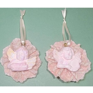 May include: Two pink angel ornaments with lace trim. Each angel is holding a scroll and has a white halo. The ornaments are tied with white ribbon.
