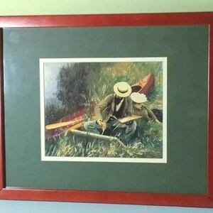 May include: Framed print of a painting depicting two people in a canoe, surrounded by tall grass. The artwork features a red canoe, oars, and figures wearing hats. The frame is a dark red color, and the matting is green.