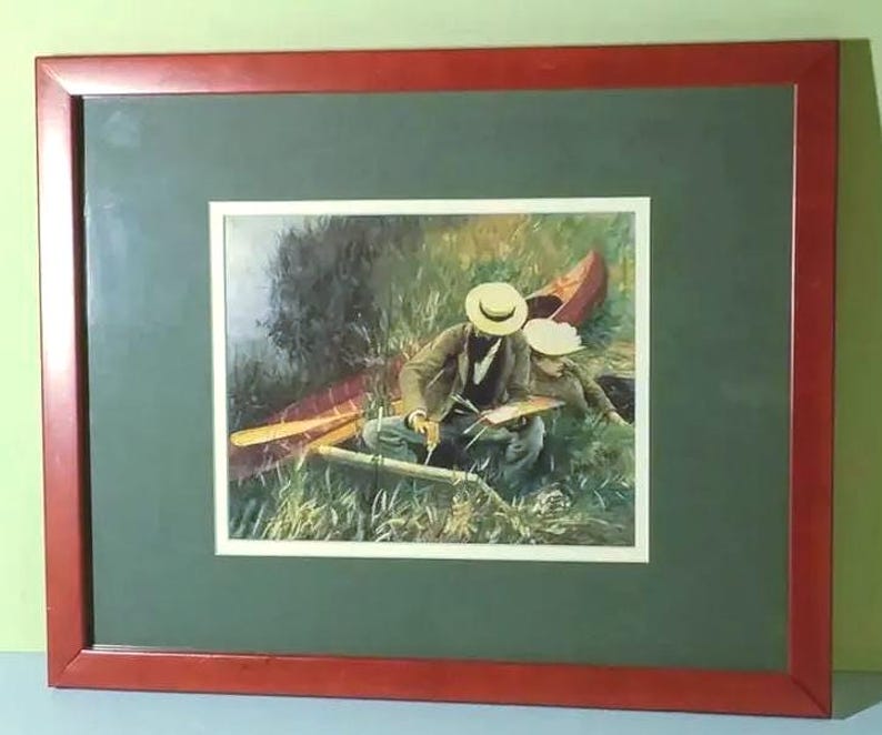May include: Framed print of a painting depicting two people in a canoe, surrounded by tall grass. The artwork features a red canoe, oars, and figures wearing hats. The frame is a dark red color, and the matting is green.