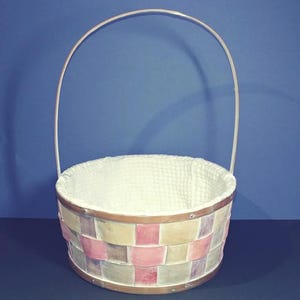 Puede incluir: Una cesta de mimbre blanca con un patrón de cuadros rosa y beige y un forro de tela blanco. La cesta tiene un asa blanca.
