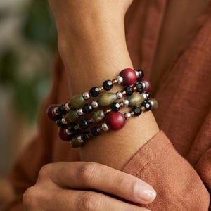May include: A multi-strand bracelet featuring a combination of round red wooden beads, olive green oval wooden beads, small black faceted beads, and metallic silver-toned spacer beads arranged in a repeating pattern around the wrist.