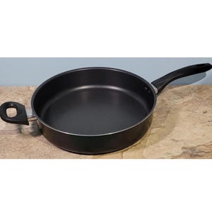 Könnte beinhalten: Eine schwarze Antihaft-Bratpfanne mit einem langen schwarzen Griff und einem kleineren seitlichen Griff. Die Pfanne steht auf einer hellbraunen Oberfläche.