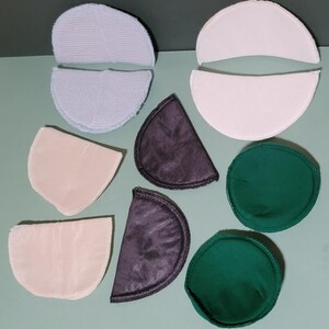 May include: A collection of assorted fabric shoulder pads in various shapes and colors, including two light blue textured pads, two white pads, two beige pads, two dark gray pads, and two circular dark green pads, all arranged on a flat surface.