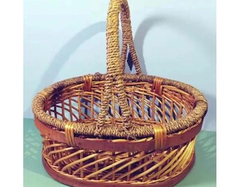 May include: Oval-shaped woven basket with a tall, arched handle. The basket is made of light brown wicker with a darker brown wooden base and trim. The handle is tightly woven, and the basket has a classic, rustic design.