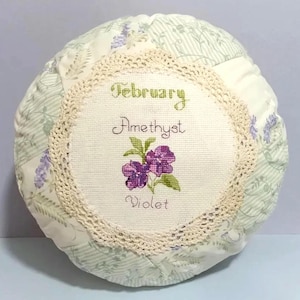 May include: A round, white pillow with a crocheted trim. The center of the pillow features a cross-stitch design with the text "February Amethyst Violet".