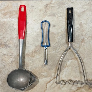 May include: Three kitchen utensils: a red ladle, a blue and silver potato peeler, and a silver potato masher with a black handle.