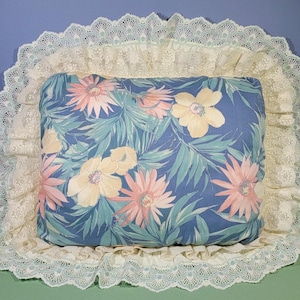 May include: A blue pillow with a floral print of pink, yellow, and green flowers. The pillow is surrounded by a white lace trim.