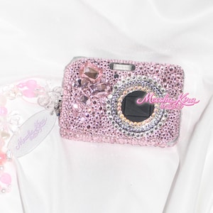 May include: Pink digital camera encrusted with rhinestones and crystals. The camera has a silver lens ring and a black lens. A pink and clear beaded wrist strap is attached to the camera. The camera has the text "Moodha Kira" on the side.