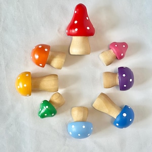 May include: A collection of eight wooden mushroom figurines in various colors, including red, orange, yellow, green, blue, purple, and pink. Each mushroom has a natural wood stem and a cap adorned with white polka dots. The mushrooms are arranged in a circular pattern.