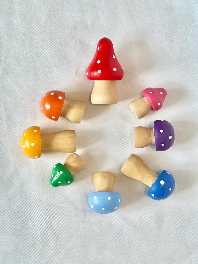 May include: A collection of eight wooden mushroom figurines in various colors, including red, orange, yellow, green, blue, purple, and pink. Each mushroom has a natural wood stem and a cap adorned with white polka dots. The mushrooms are arranged in a circular pattern.