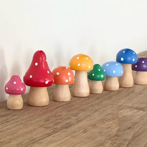May include: A collection of seven wooden mushroom figurines in various colors, including pink, red, orange, green, light blue, dark blue, and purple. Each mushroom has a natural wood stem and white polka dots on the cap.