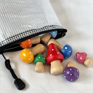 May include: A collection of colorful wooden mushroom toys with white polka dots spills from a white mesh zippered bag. The mushrooms have red, blue, green, orange, yellow, pink, and purple caps. The stems are natural wood.
