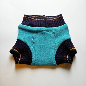 Puede incluir: Un pañal para mascotas de color turquesa y azul marino con un diseño de punto. El pañal presenta una almohadilla absorbente turquesa y puños azul marino con rayas rojas y doradas. El producto está diseñado para mascotas.